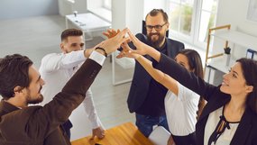 Dein Geld. Deine Zukunft. Mehrere Personen stehen um einen Tisch und legen ihre Hände in der Mitte übereinander. Sie tragen Businesskleidung und wirken konzentriert. Im Hintergrund sind große Fenster und helle Büroräume zu sehen.